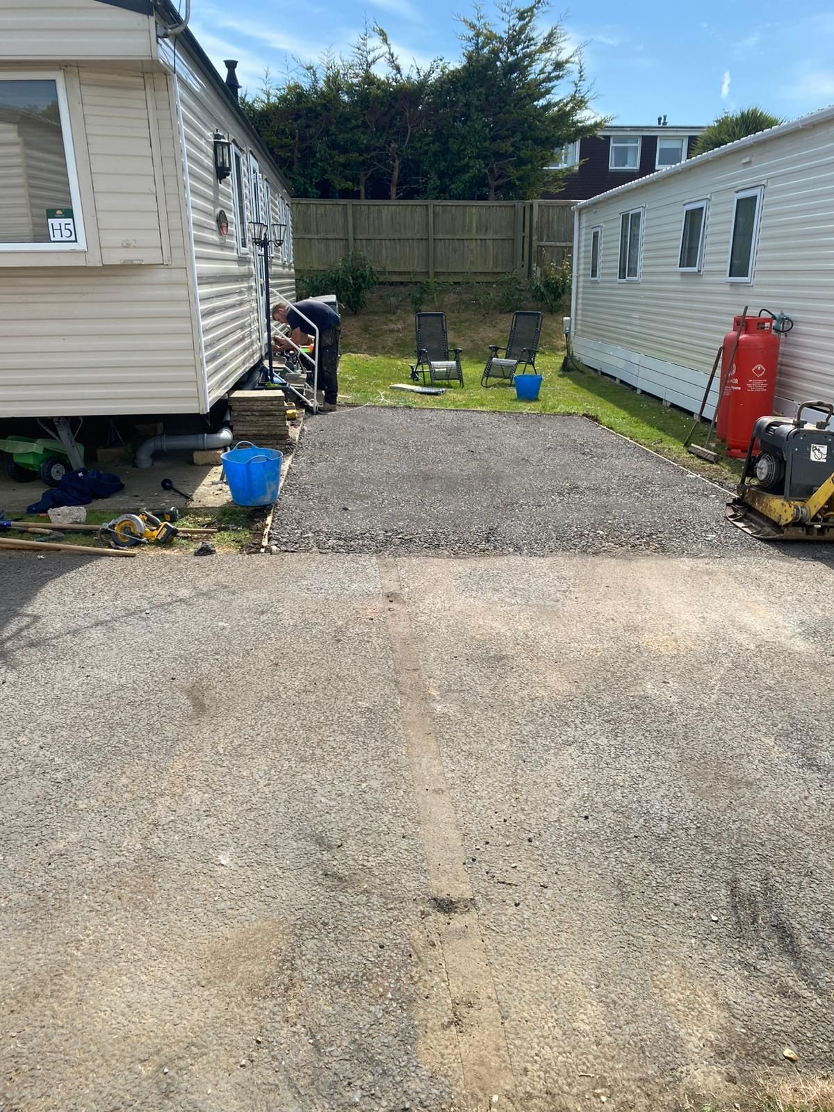 Gravel driveway between two mobile homes; person works on ground, chairs sit on grass, blue sky.