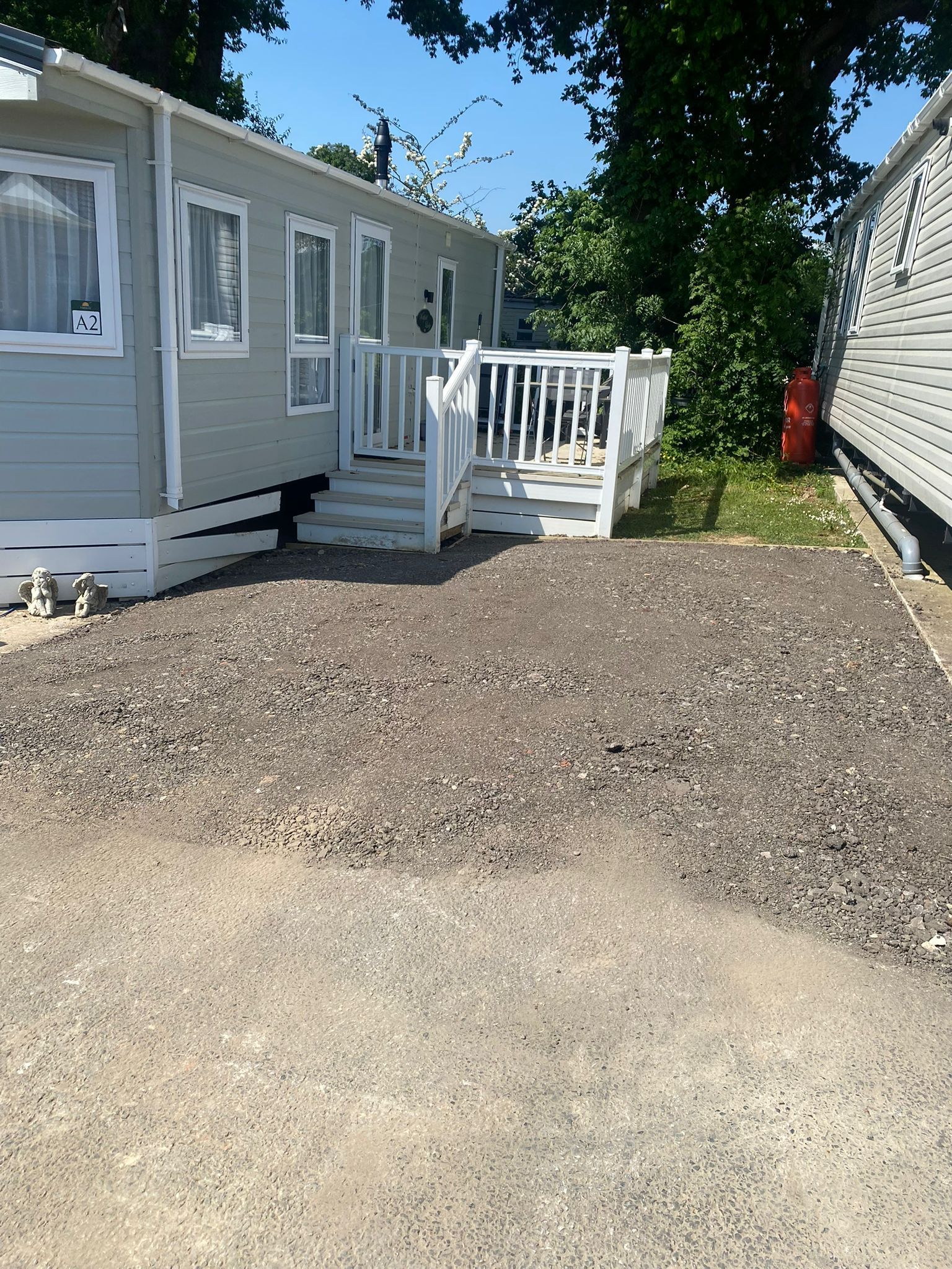 Gray mobile home with white deck, gravel parking area, and trees.