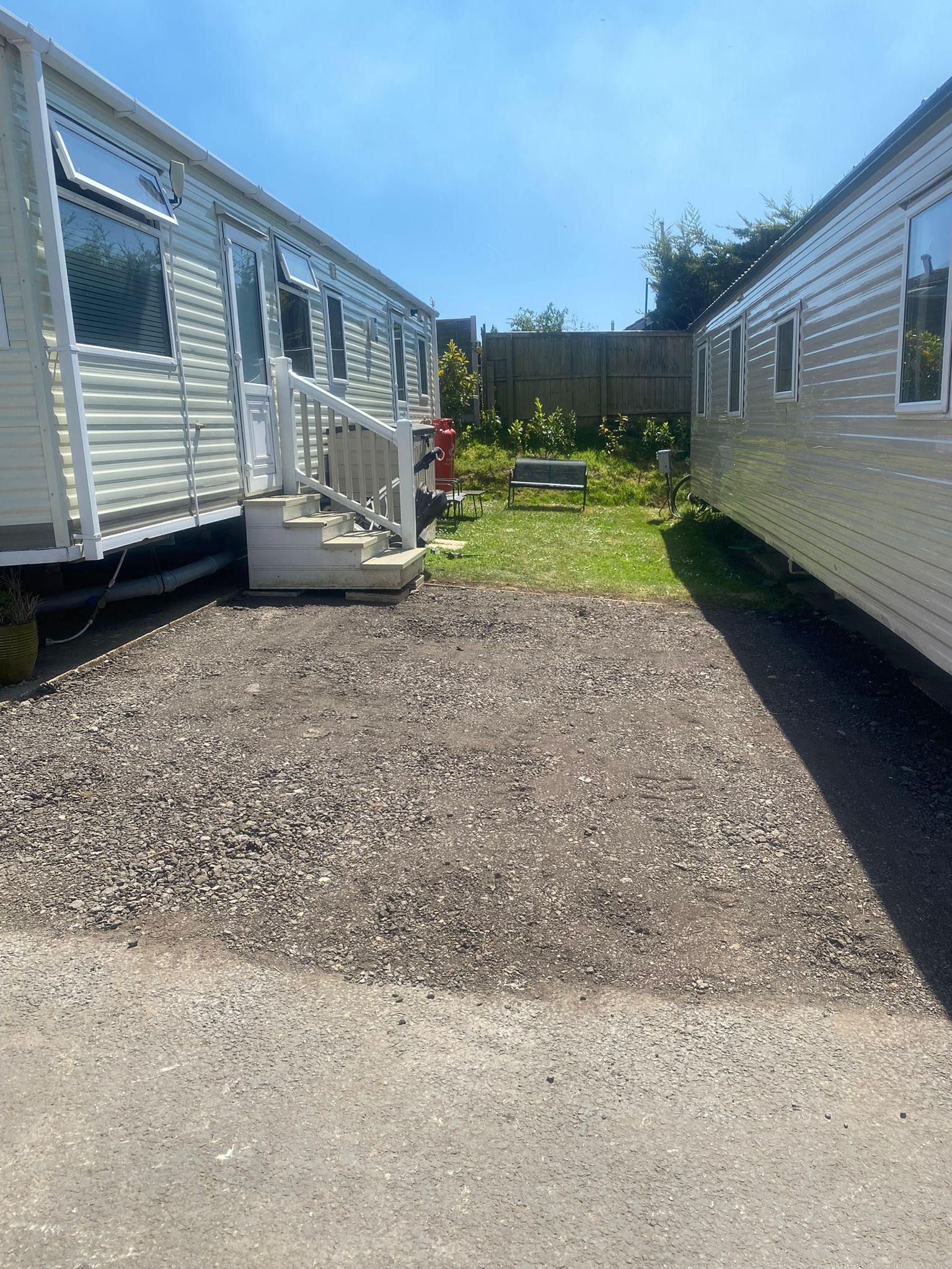 Gravel parking area between two light-colored mobile homes, with small grassy area in the back on a sunny day.