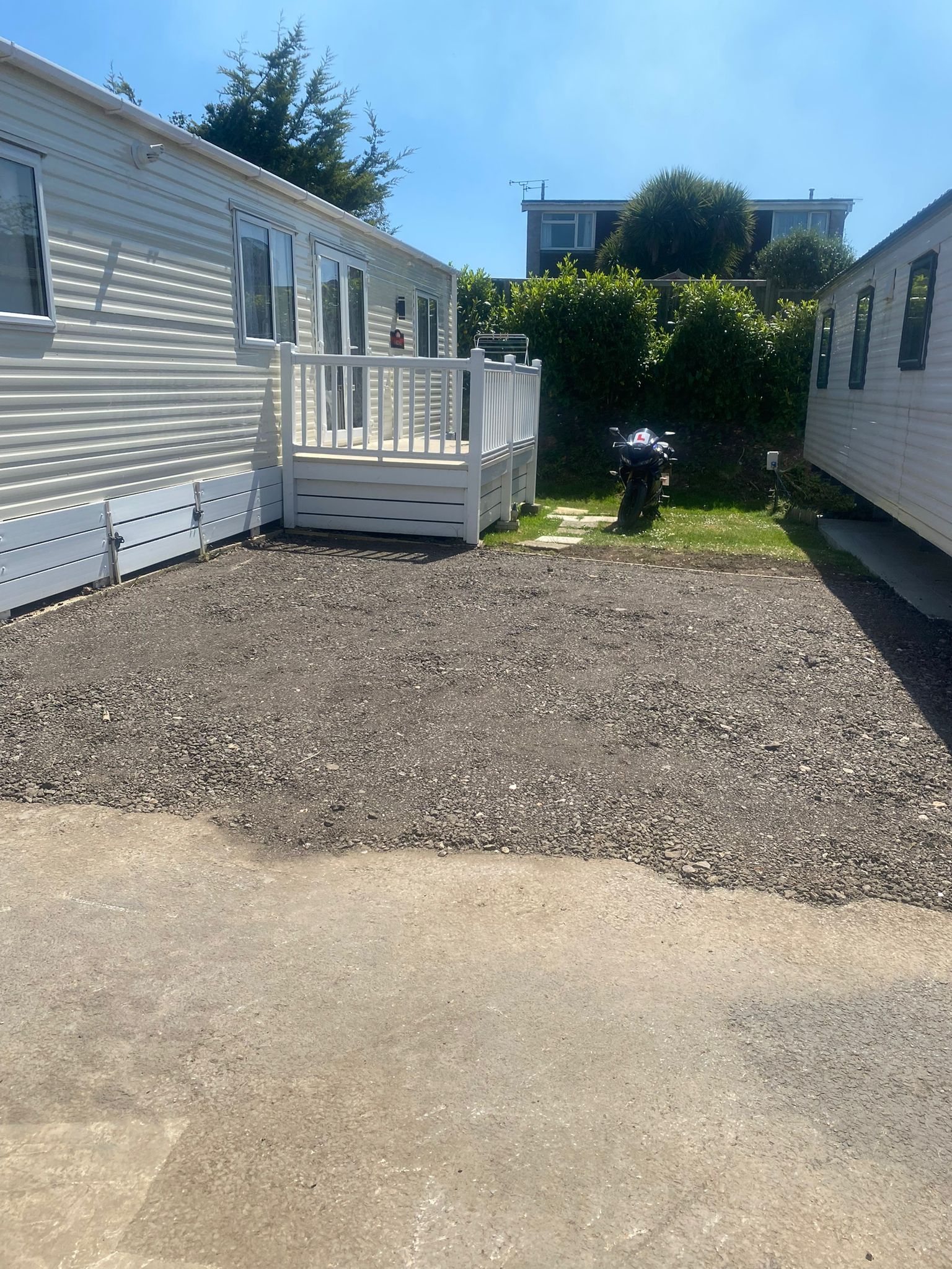 Two white mobile homes, gravel patio, deck, lawn, grill, bright blue sky.