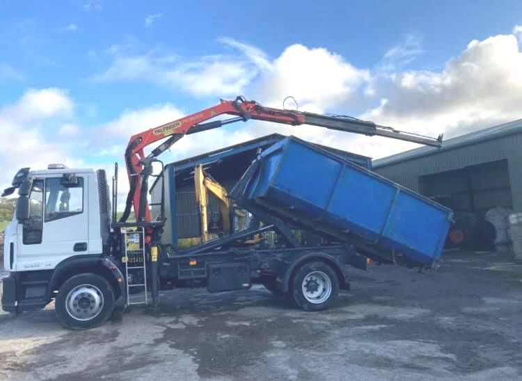 A dump truck with a crane attached to it is being loaded with a blue dumpster.