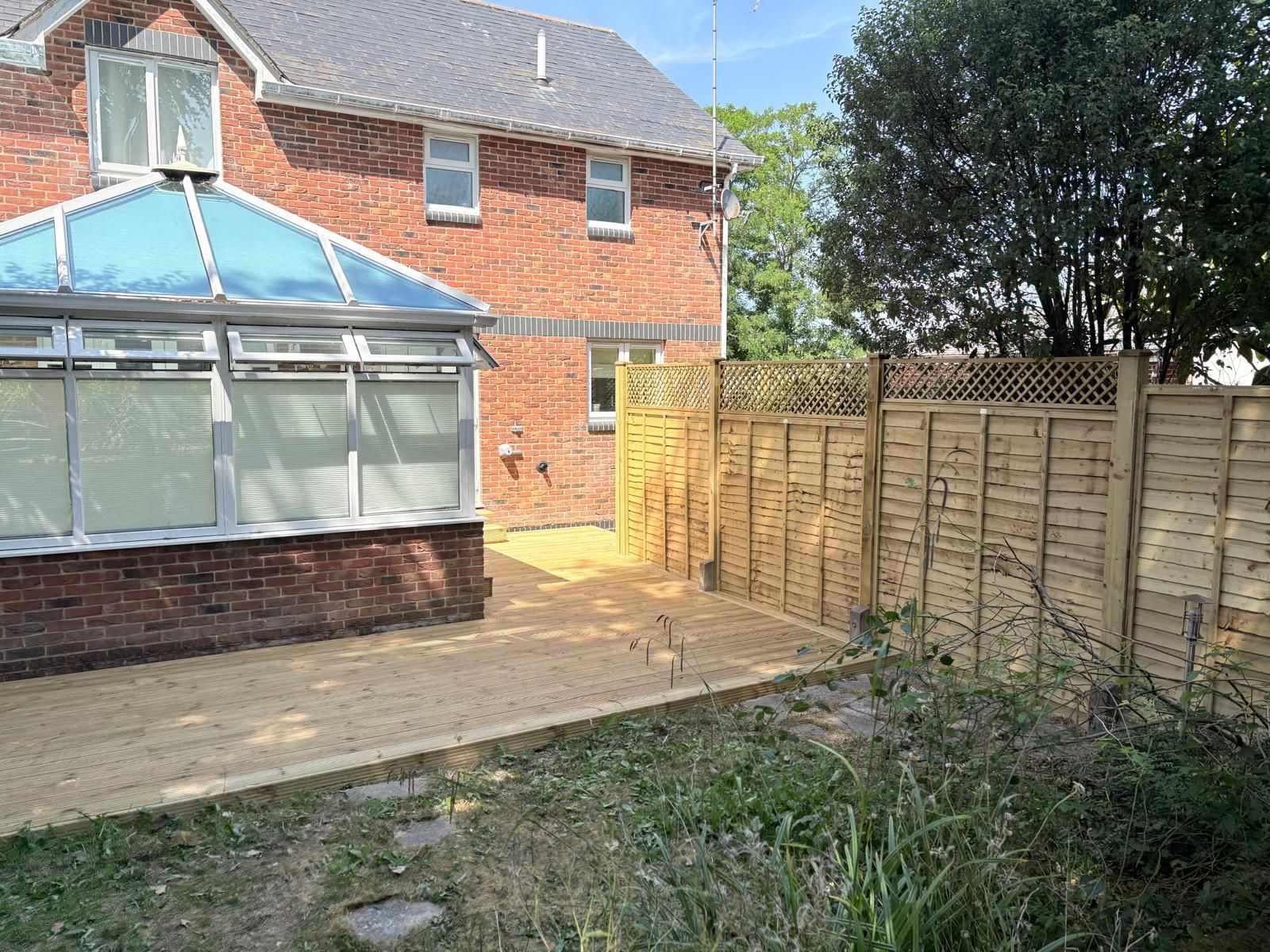Backyard patio beside a brick house with a conservatory and wooden fence, bordered by trees and grass.