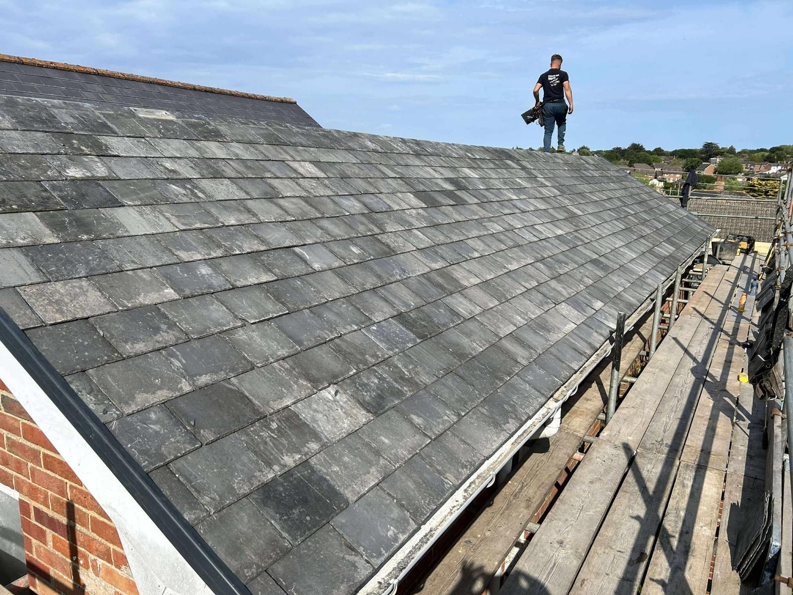 Person on a gray slate roof, construction scaffolding below. Brick building, blue sky.
