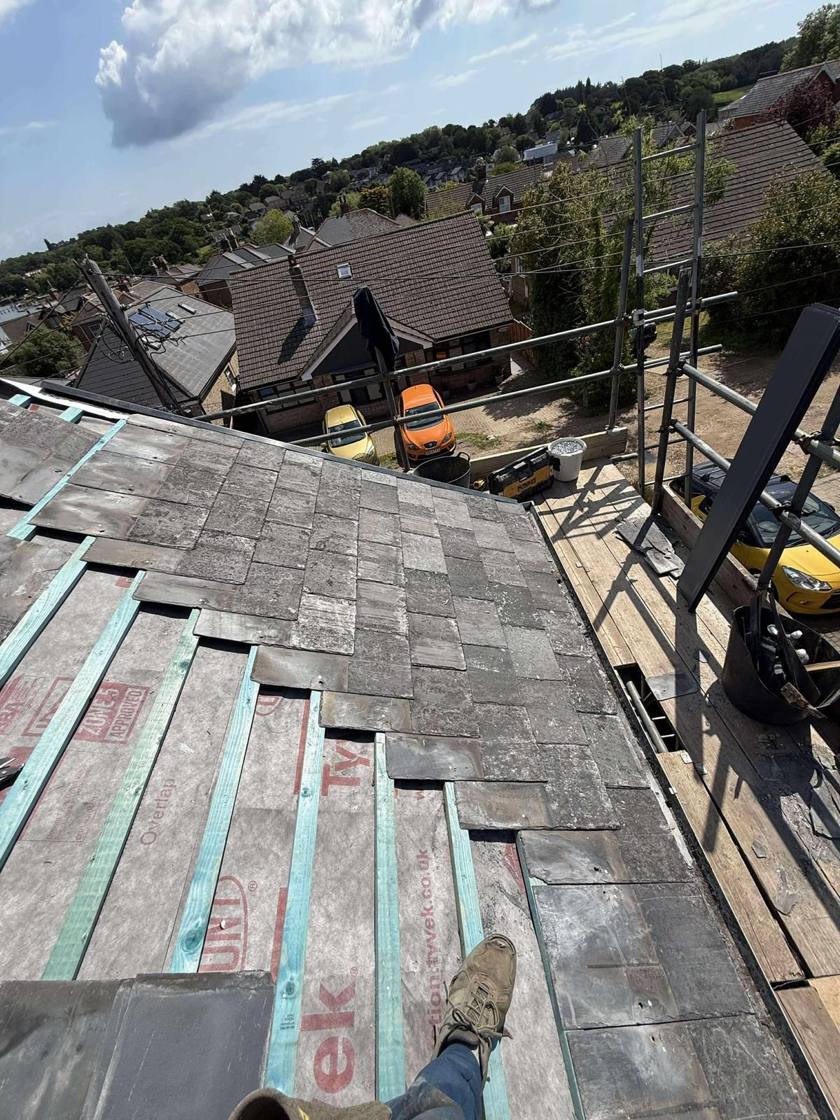Roofer on a slate roof, partly installed. Scaffolding nearby, houses and cars in background, sunny day.