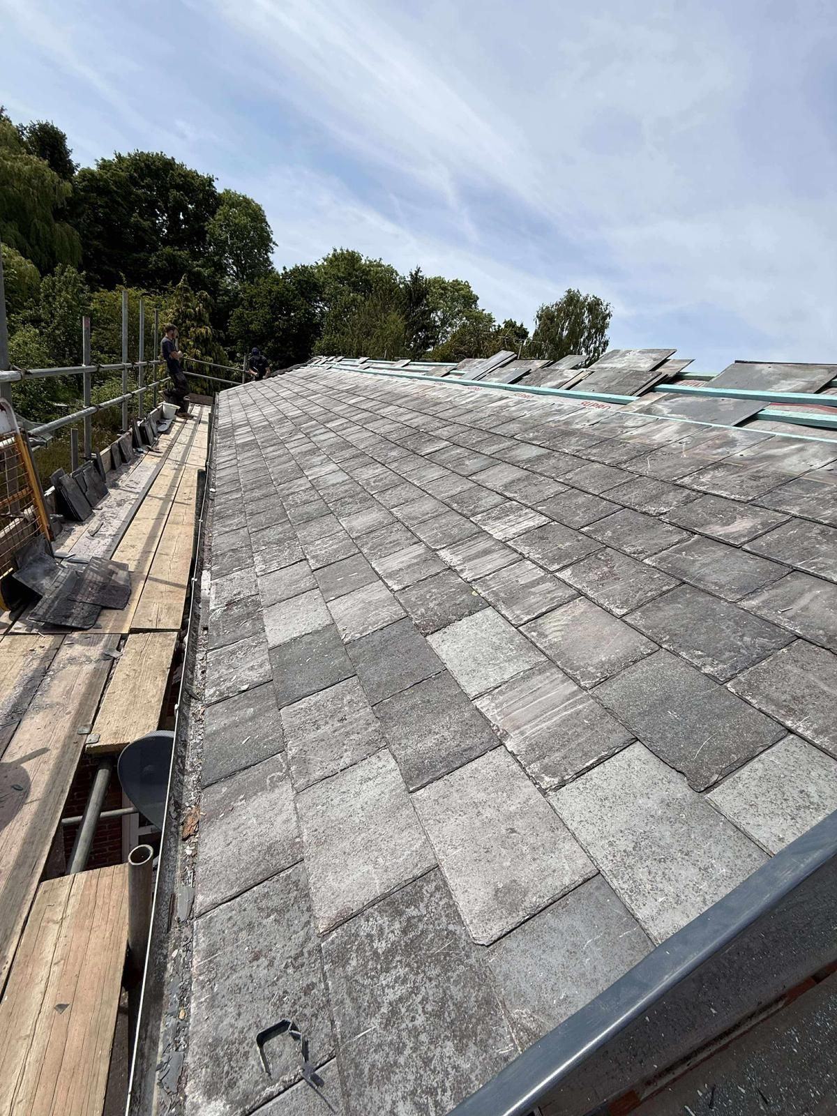 Rooftop with gray shingles, scaffolding, and trees under a bright blue sky.