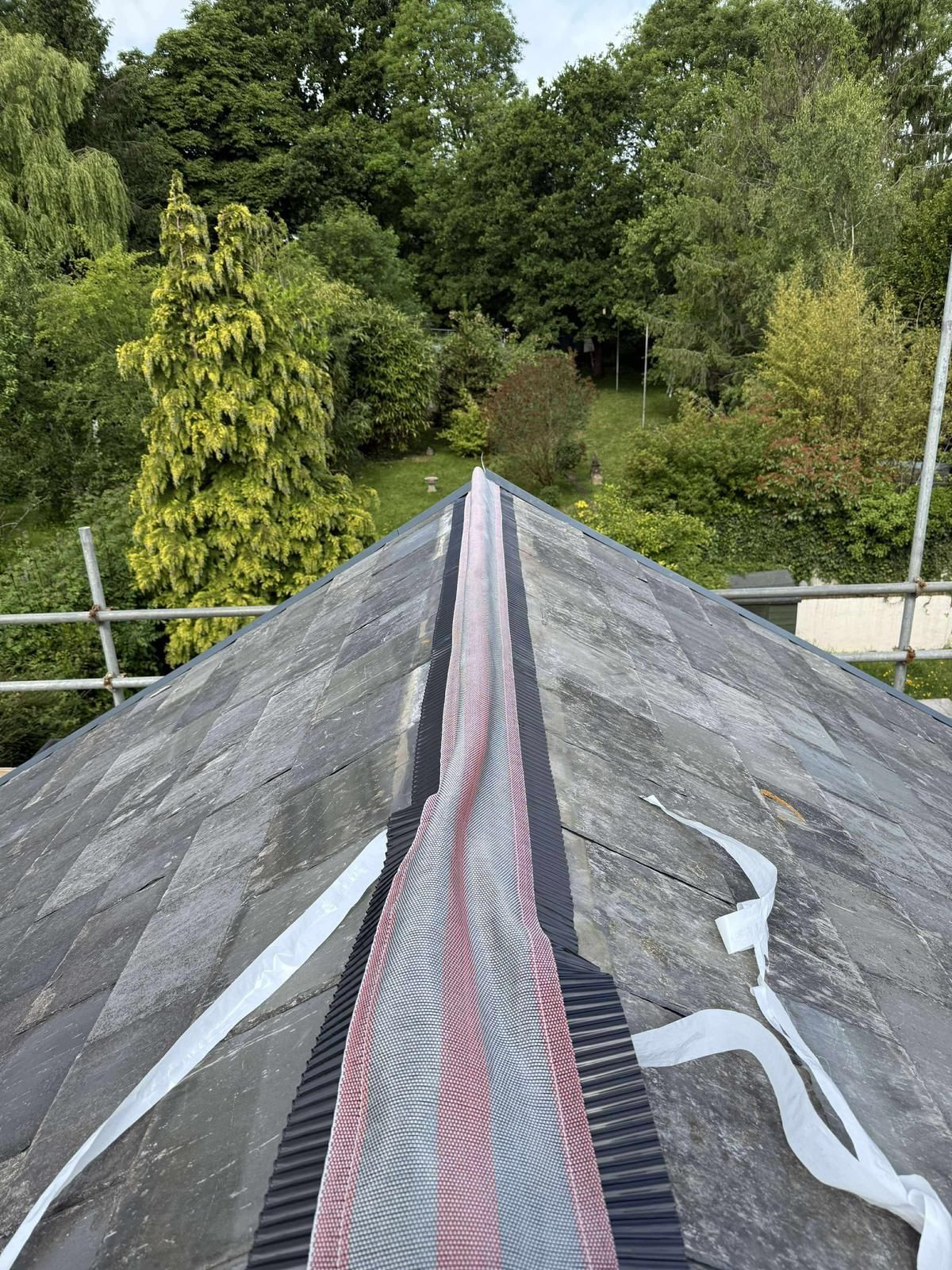 Roof with black slate tiles and a strip of red and black flashing. Green trees in the background.
