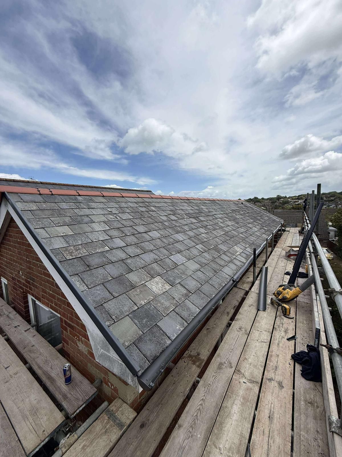 A brick building with a slate roof under a cloudy sky. Scaffolding runs along the roof edge.