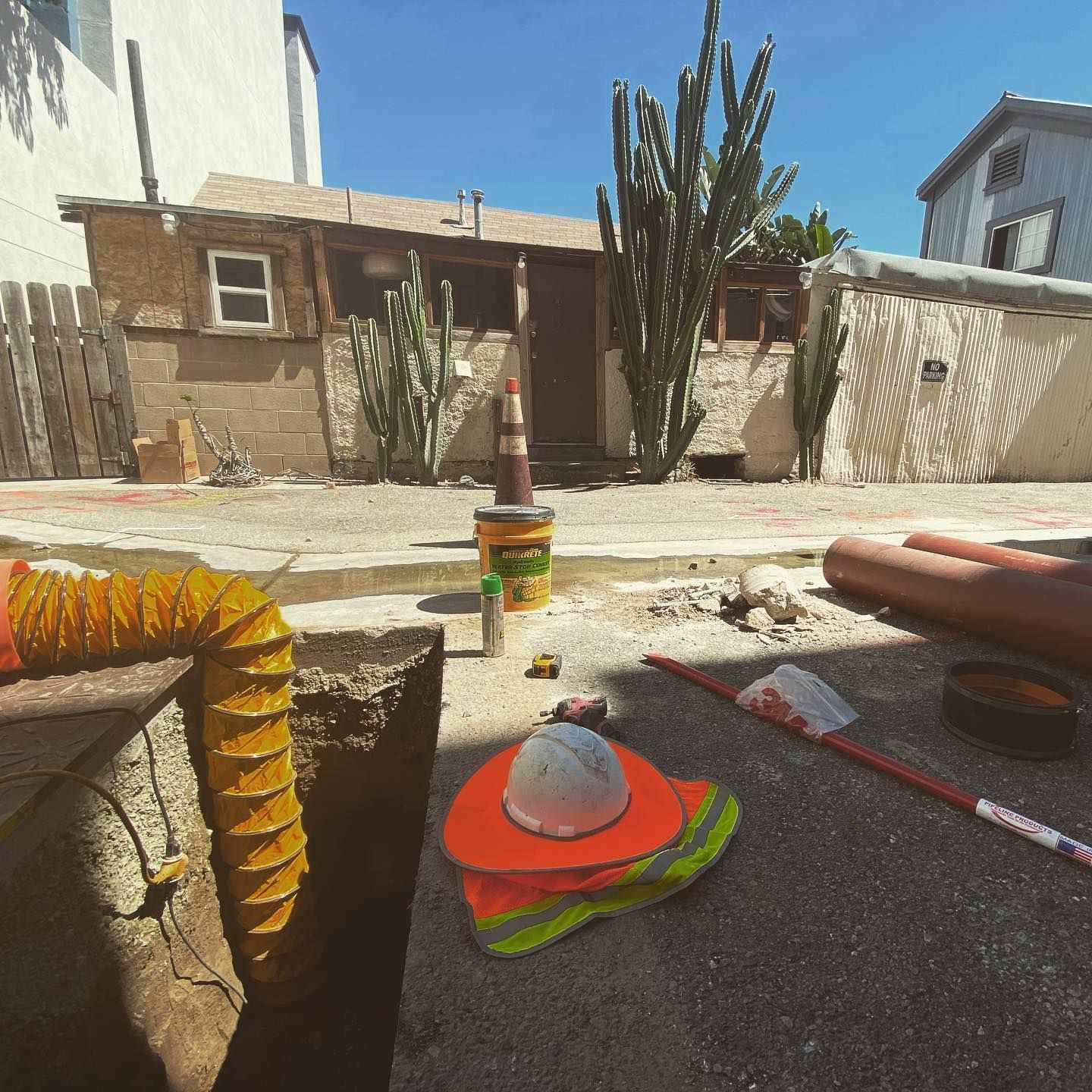 Construction site with hard hat, orange vest, and cactus in front of a small building.