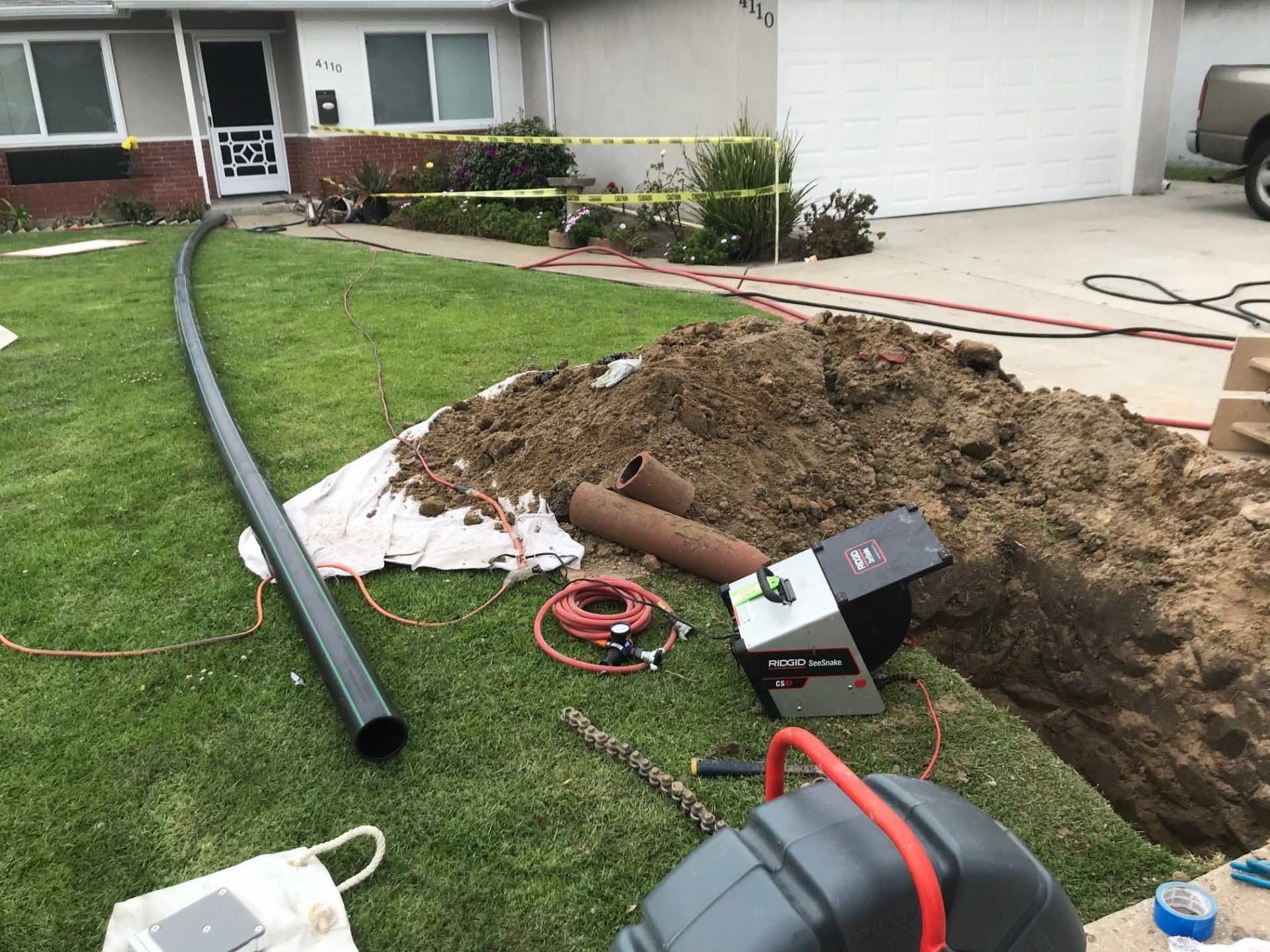 A trench dug in a lawn with equipment for underground pipe work. A black pipe extends across the grass.