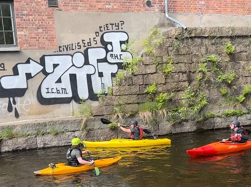 En gruppe mennesker kajakkpadler ned en elv foran en murvegg med graffiti på.