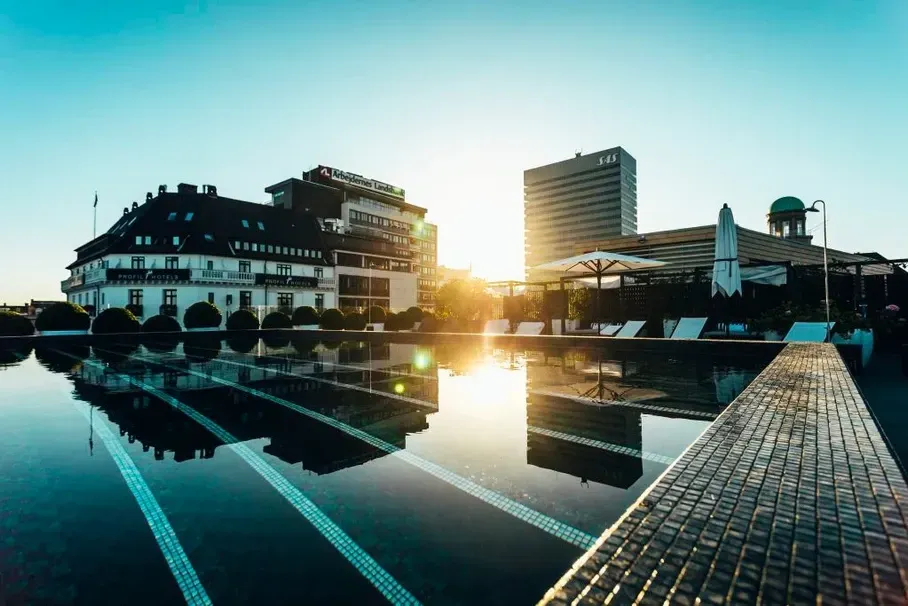 Et stort svømmebasseng med en bygning i bakgrunnen og solen reflektert i vannet.