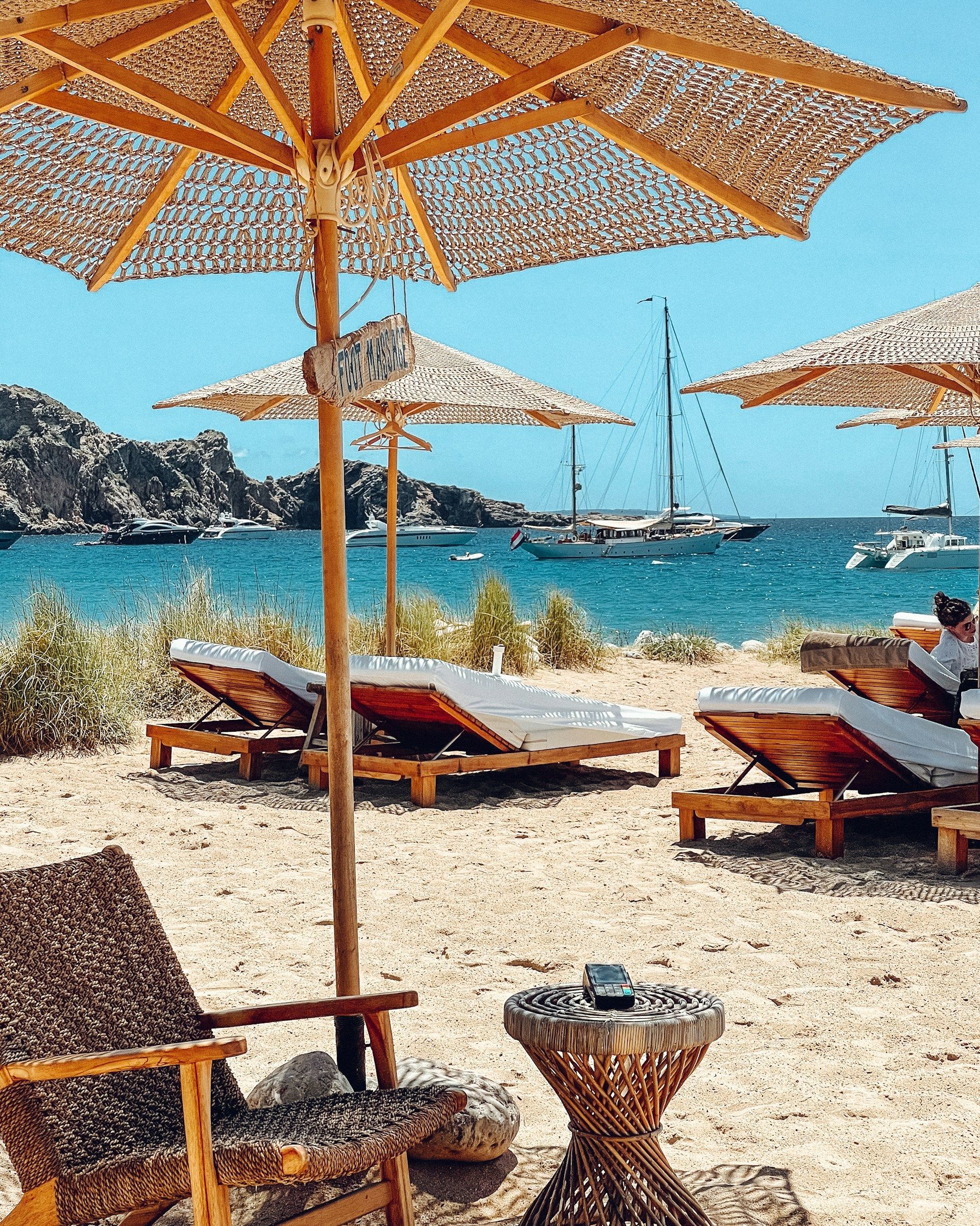En strand med stoler og parasoller og båter i bakgrunnen