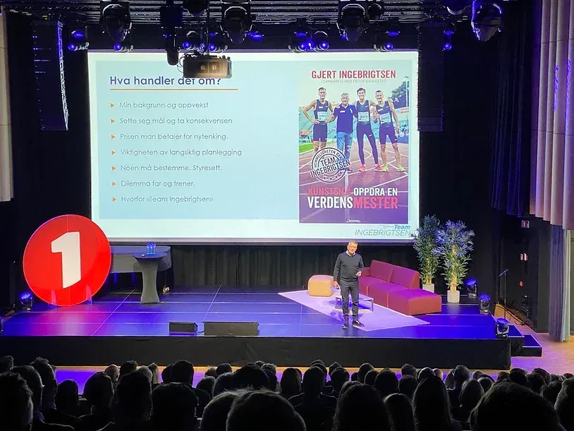 En mann holder en presentasjon på en scene foran en stor skjerm.