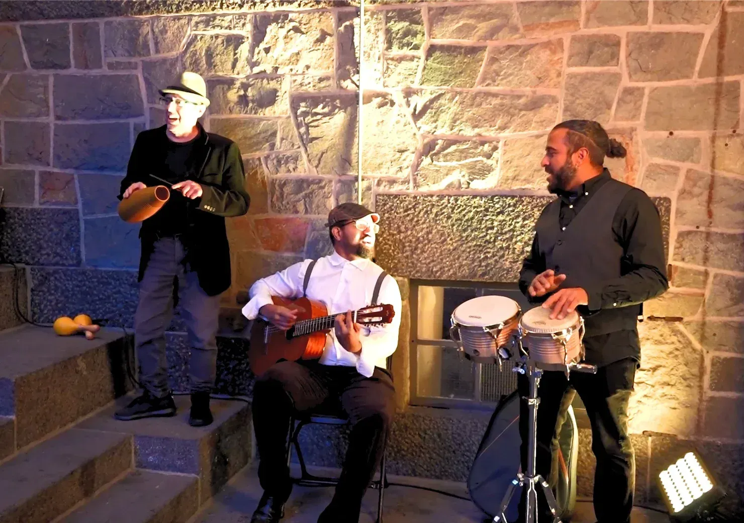Tre menn spiller instrumenter foran en steinmur