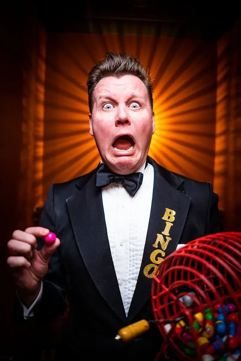 En mann i smoking holder en bingoball og lager et morsomt ansikt.