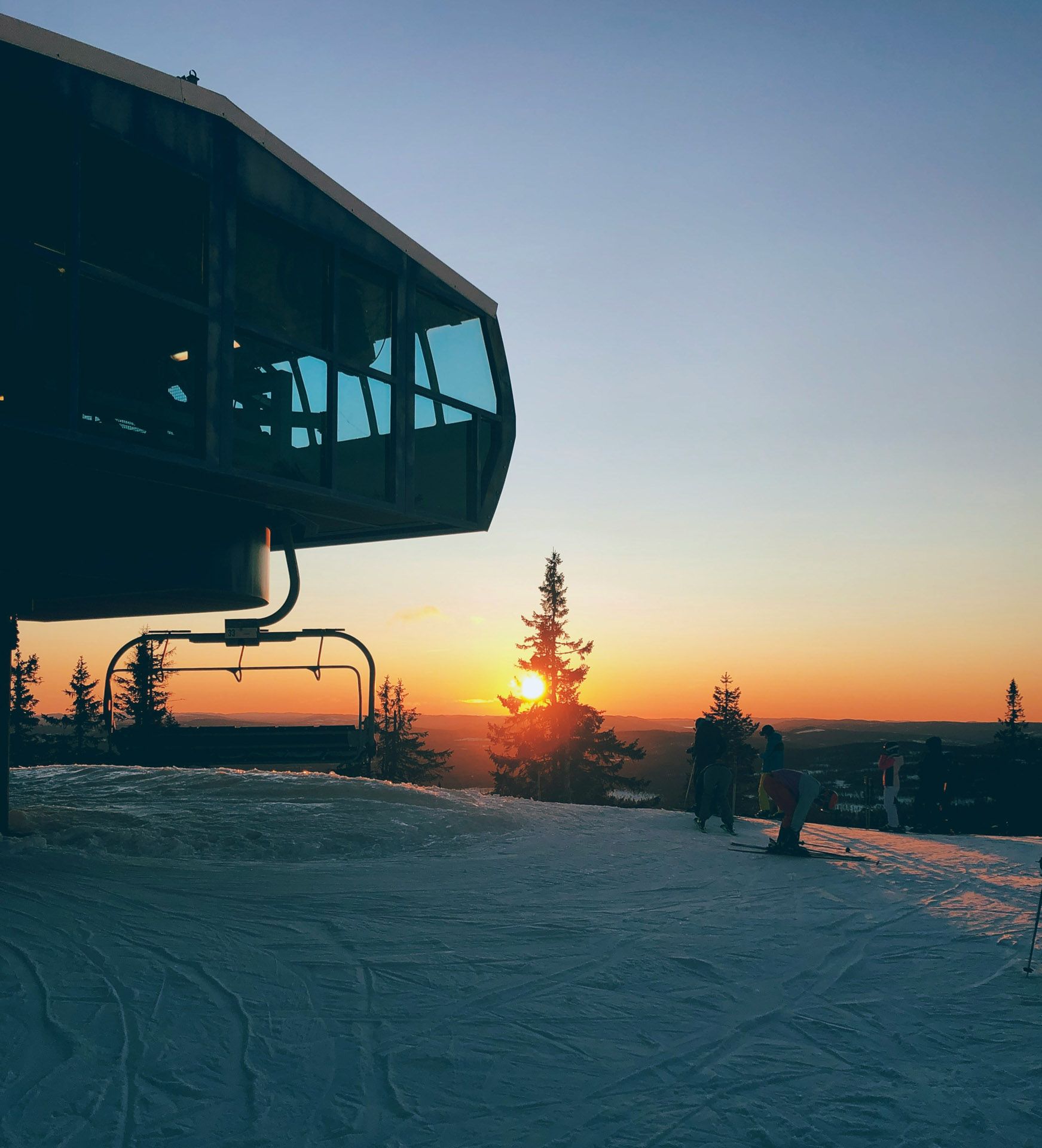 Skiheisstasjon ved solnedgang, med utsikt over en snødekt bakke og trær; oransje og blå himmel.