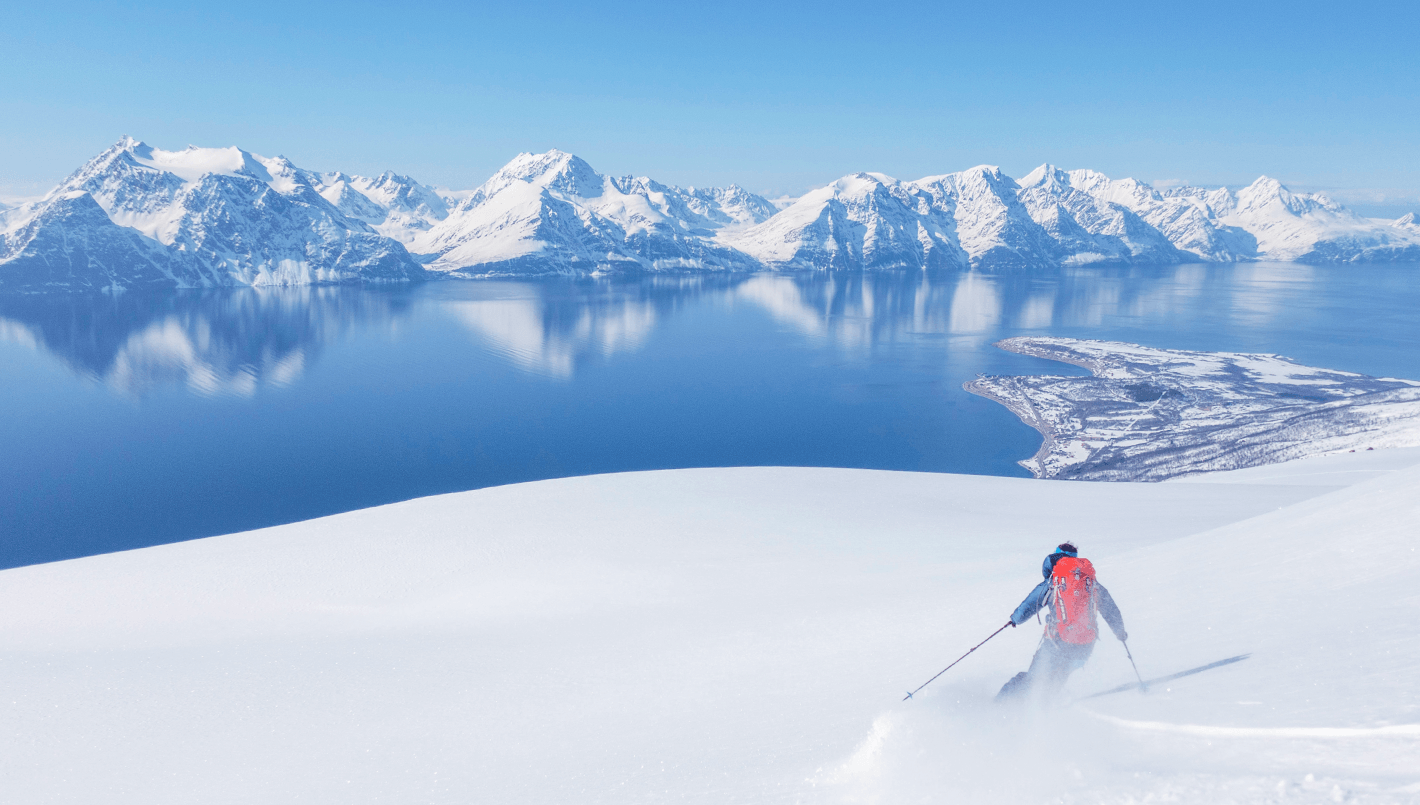 En person går på ski ned en snødekket skråning med fjell i bakgrunnen.