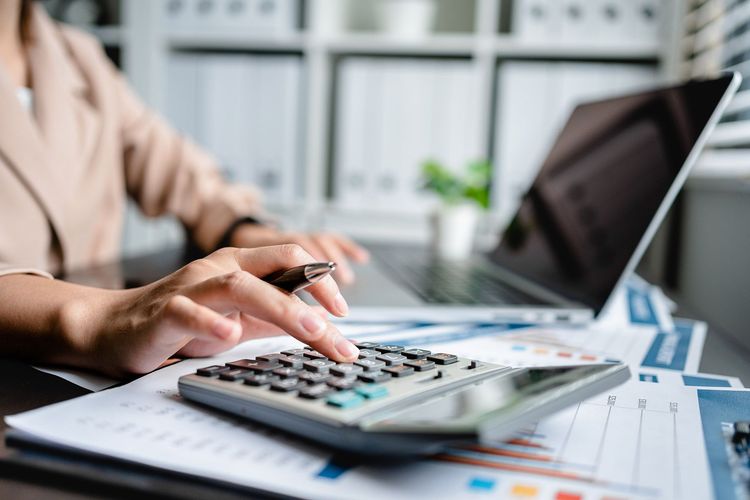 Hands calculating bills on a calculator beside financial charts and a laptop on a desk