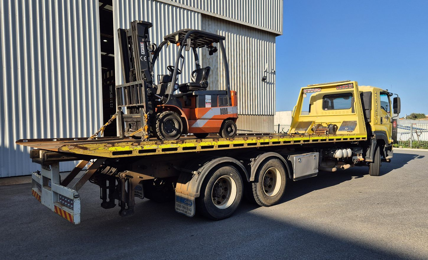 Balticore tilt tray truck with a forklift chained on outside a warehouse