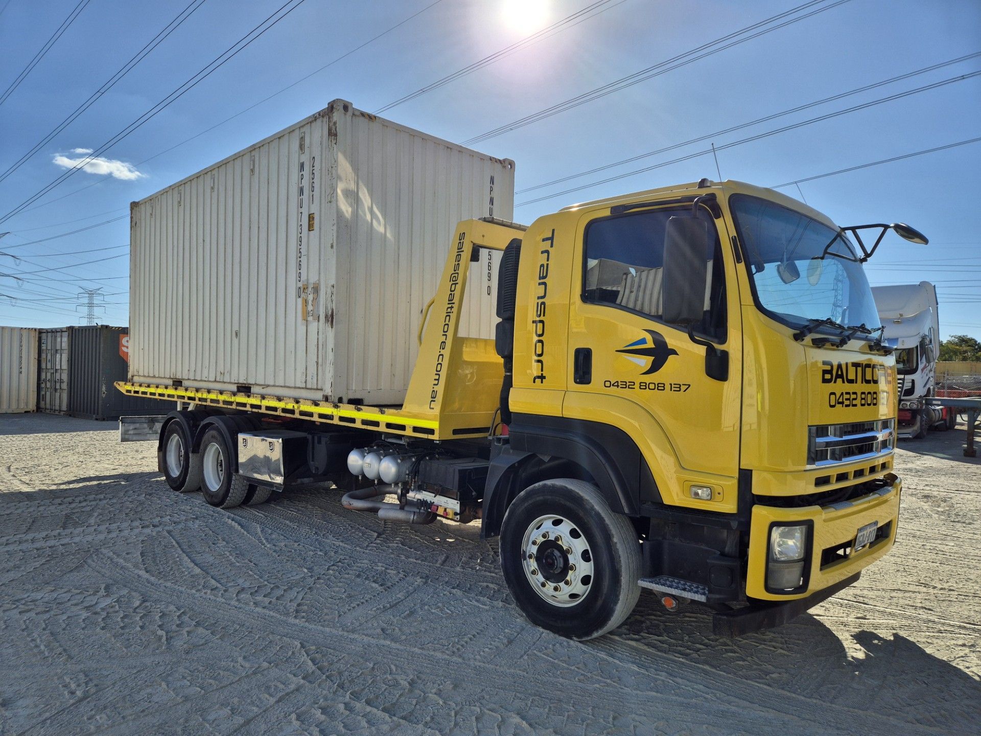 Yellow flatbed truck carrying a white shipping container in a yard with other containers under a blue sky.