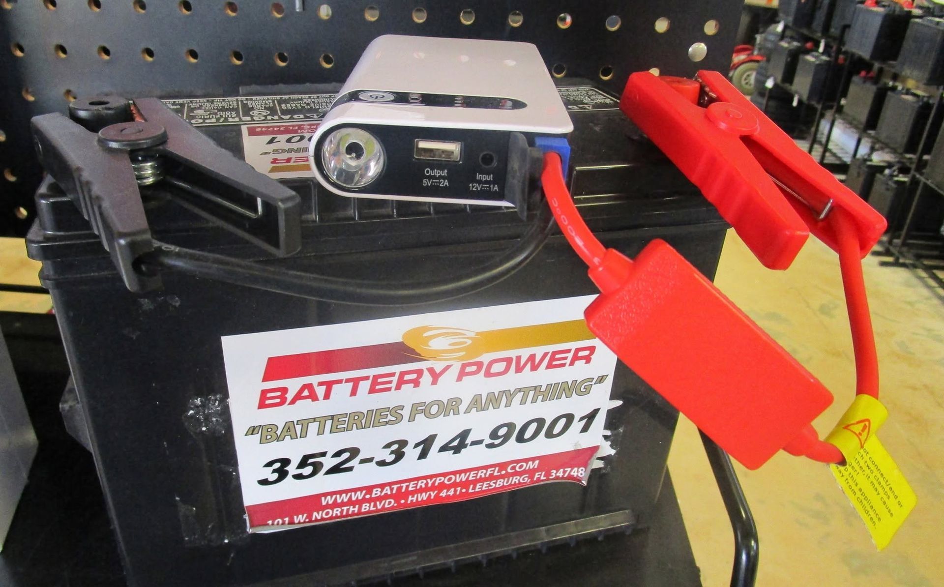 Black battery with red jumper clamps and a battery charger on a store shelf, labeled “Battery Power.”