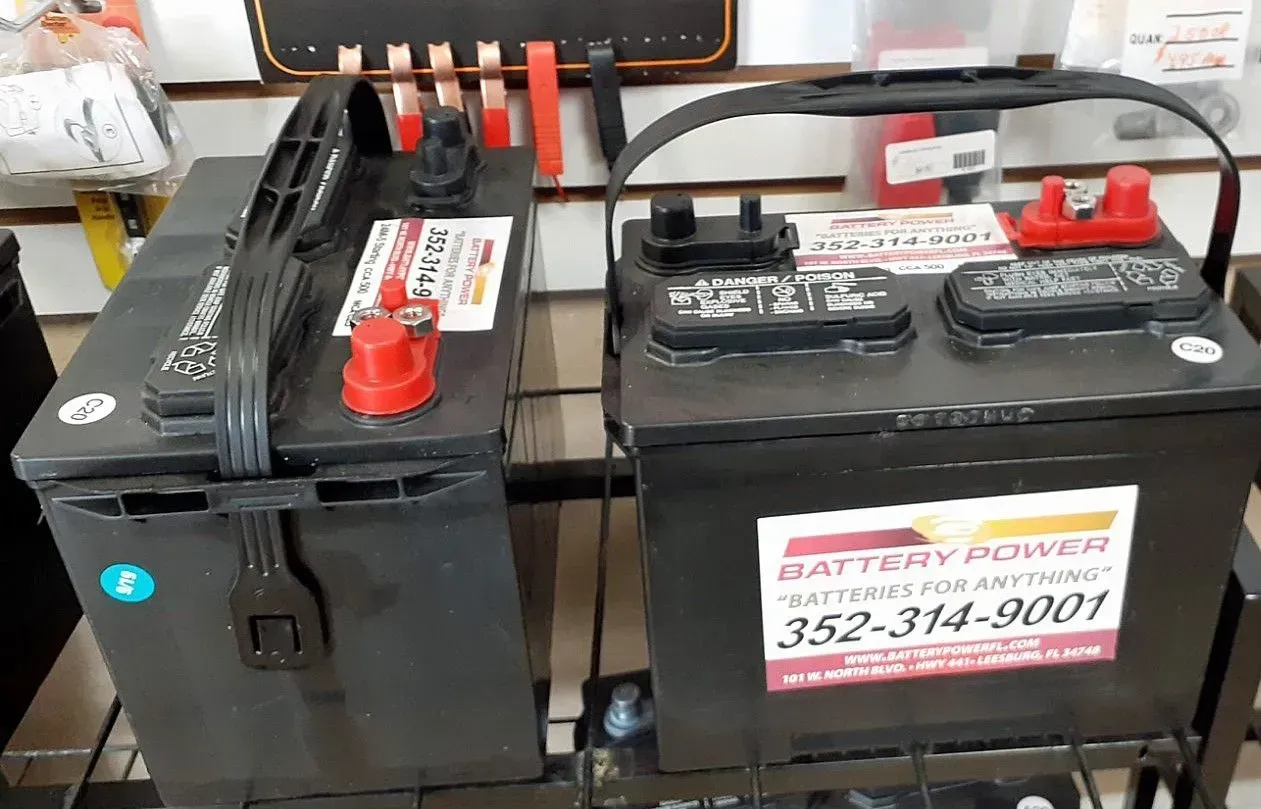 Two black car batteries with red terminals on a shelf, one labeled with a battery service sign.