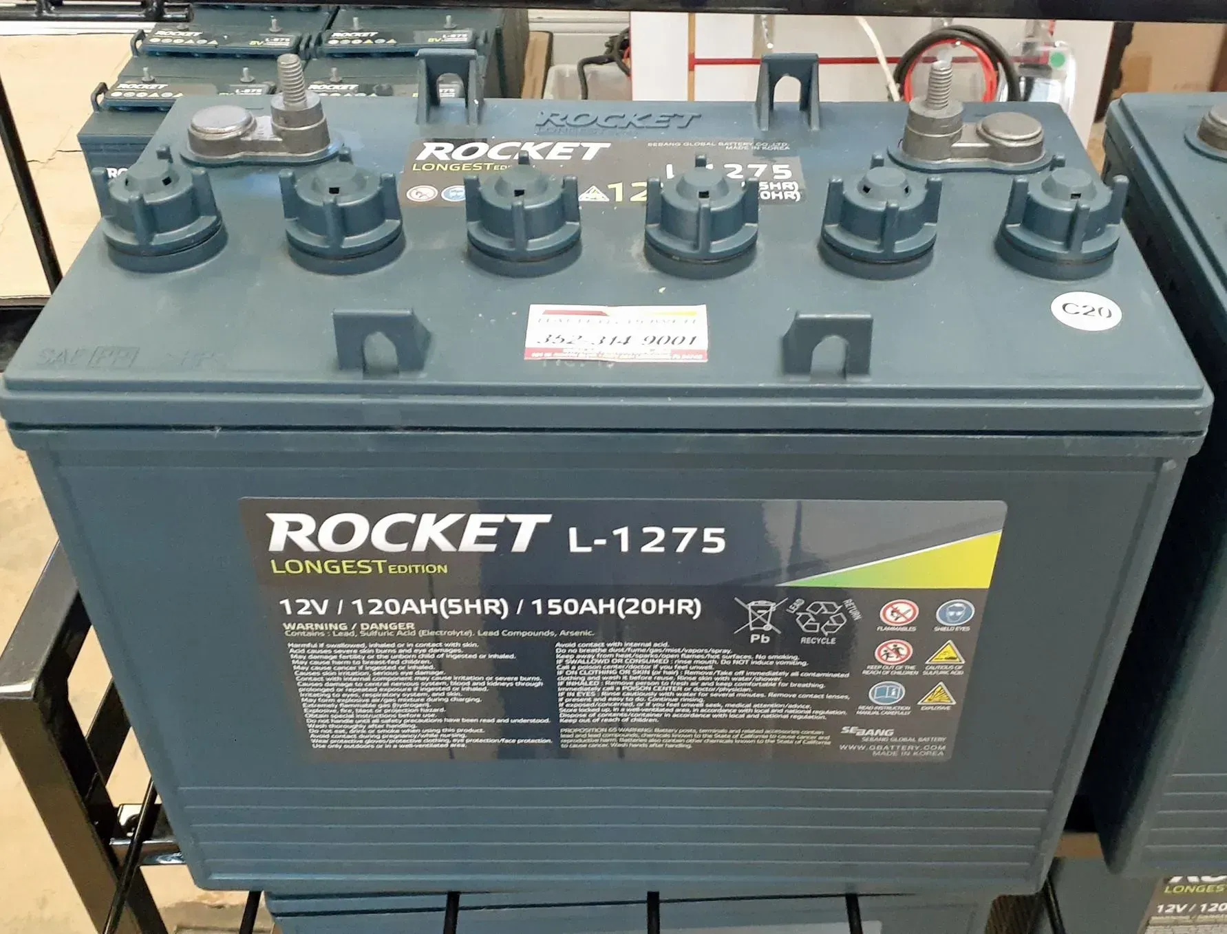 Gray Rocket L-1275 car battery with six caps, shown on a shop floor