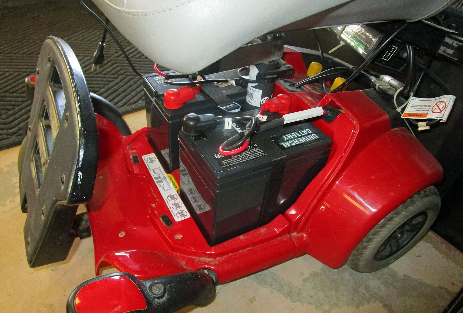Red mobility scooter with black seat and control panel, parked indoors with a large white object behind it