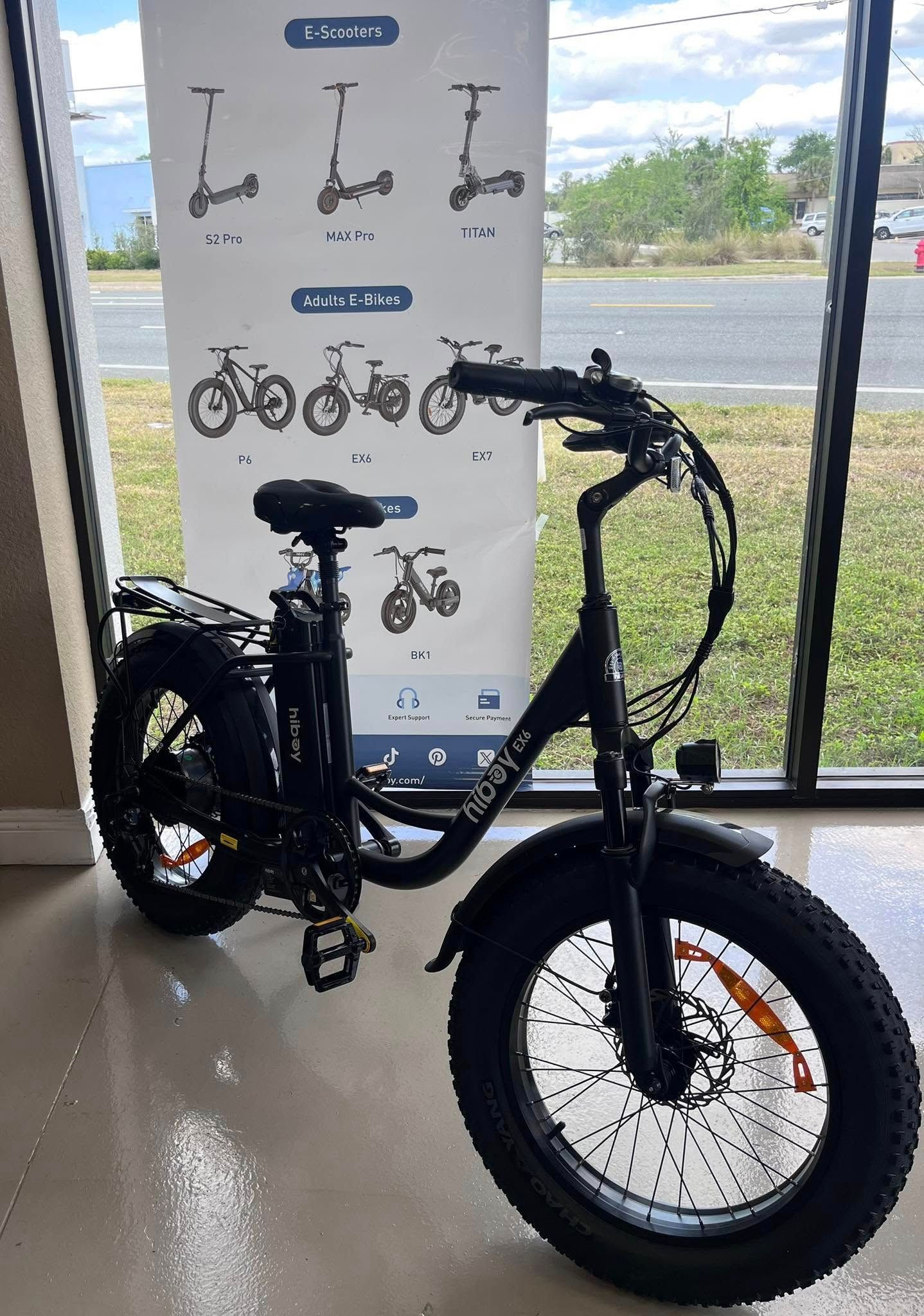 Black fat-tire electric bike indoors by a window, with a model display poster behind it.