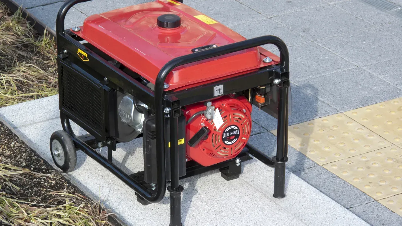 Red portable generator with black frame and wheels on a sidewalk near tactile paving.