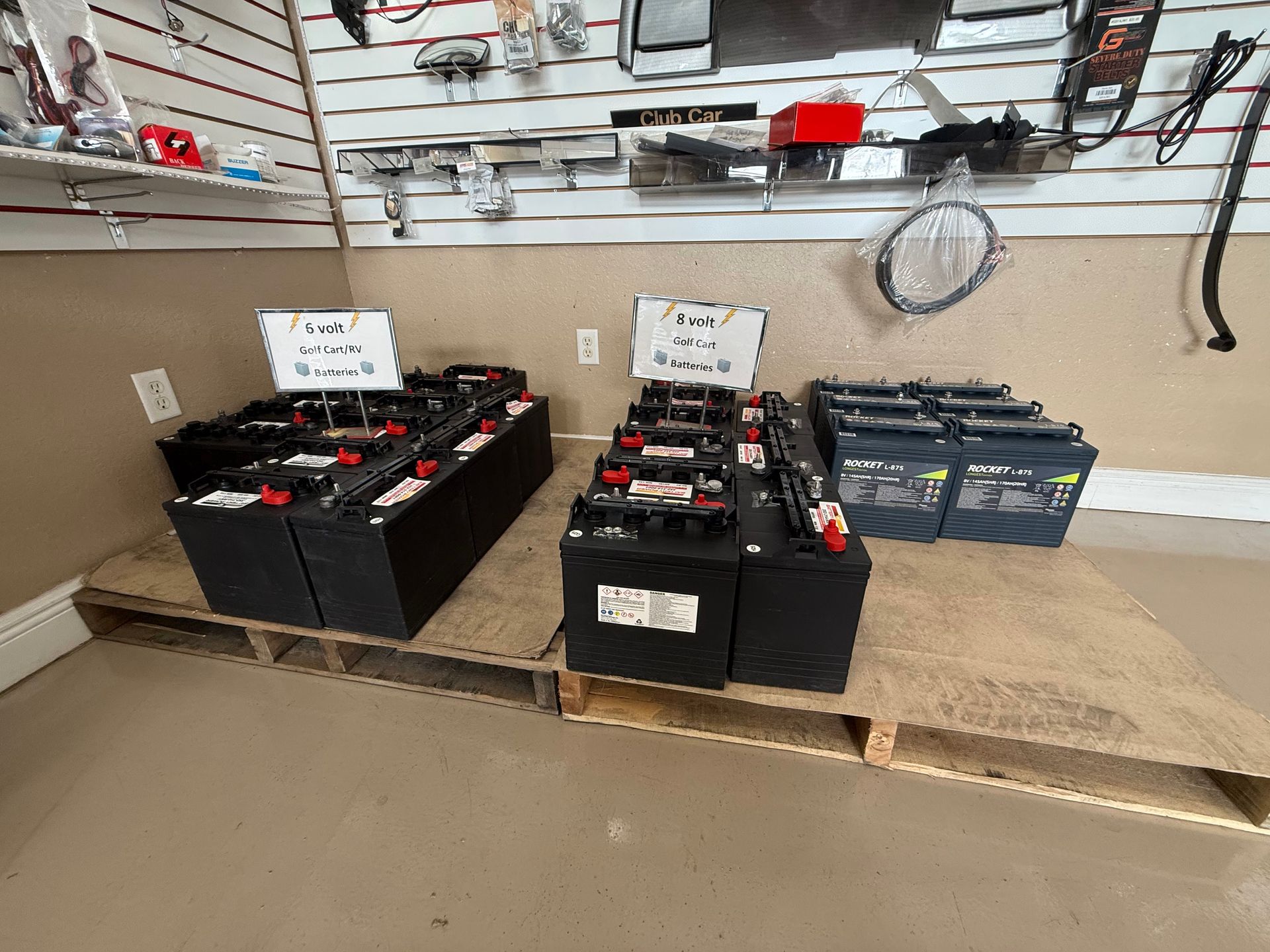 Black batteries lined on a wooden pallet in a workshop, with tools hanging on the wall behind them.