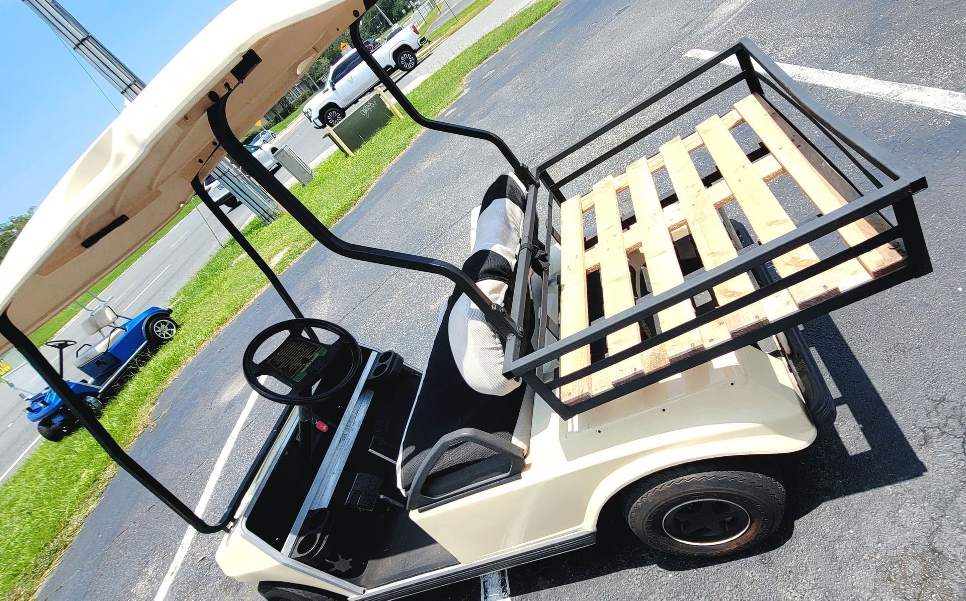 White golf cart parked on a paved path beside a grassy field, with a rear cargo rack and roof canopy.