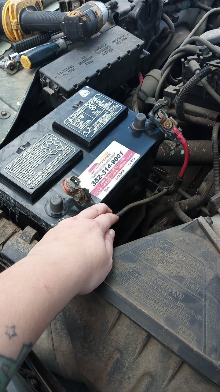 Hand pointing at a car battery under the hood, with wiring and engine components visible.