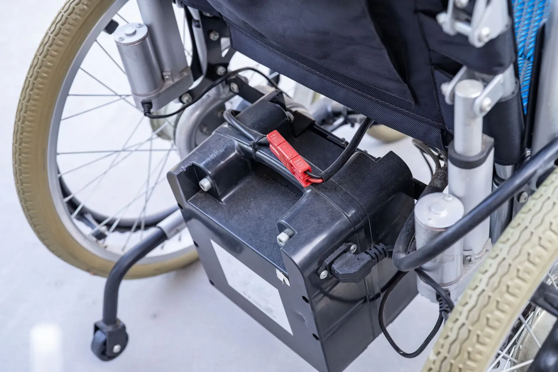 Close-up of a wheelchair battery pack mounted beneath the seat, with wiring and rear wheels visible.
