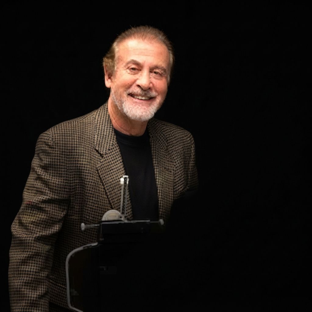 Smiling man in a dark jacket standing against a black background, speaking at a microphone