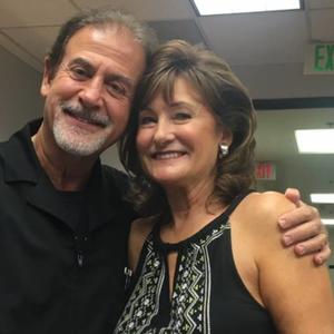 Two smiling people pose indoors, one with an arm around the other, under fluorescent lights.