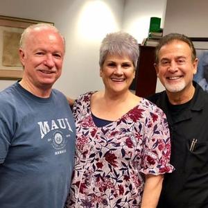 Three smiling people standing indoors, with a floral blouse and two casual shirts.