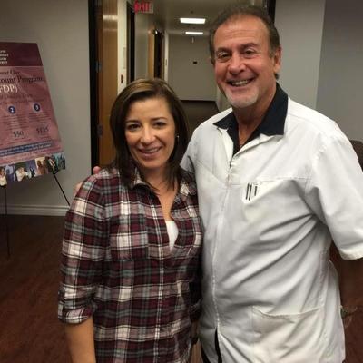 Two smiling people standing indoors, one in a white coat and one in a plaid shirt