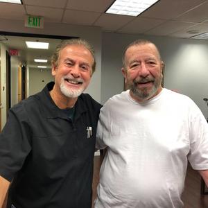 Two men smiling indoors in a hallway, one in a black shirt and one in a white shirt.