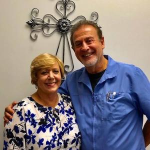 Two smiling adults standing together indoors, with a decorative wall cross behind them.