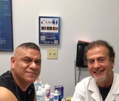 Two men smiling in a clinic office, with medical supplies and a wall sign behind them.