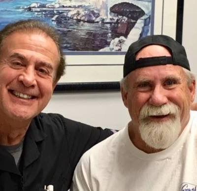 Two smiling men pose indoors in front of a framed wall art piece.