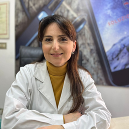 Donna in camice bianco seduta a una scrivania, sorridente in un ufficio con un'opera d'arte astratta alle pareti.