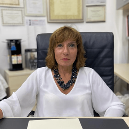 Donna seduta a una scrivania in un ufficio, con indosso una camicetta bianca e una collana scura, con certificati incorniciati alle sue spalle.