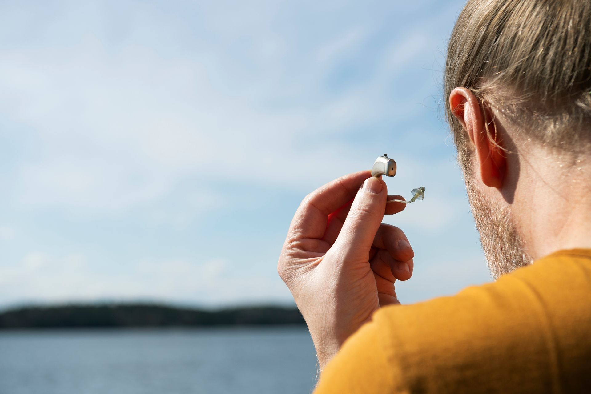 Una persona tiene un auricolare vicino all'orecchio mentre guarda un lago tranquillo sotto un cielo azzurro.
