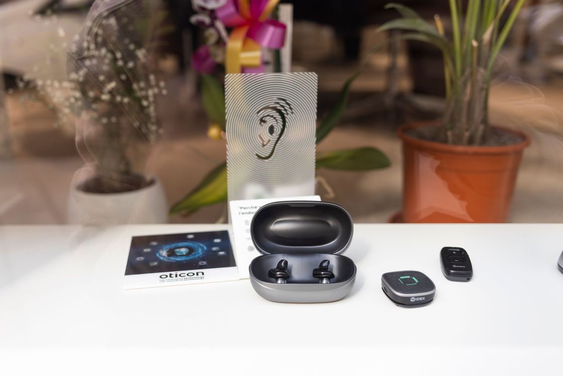 Auricolari wireless nella custodia di ricarica con telecomando, scheda e pianta sullo sfondo, su un tavolo bianco.