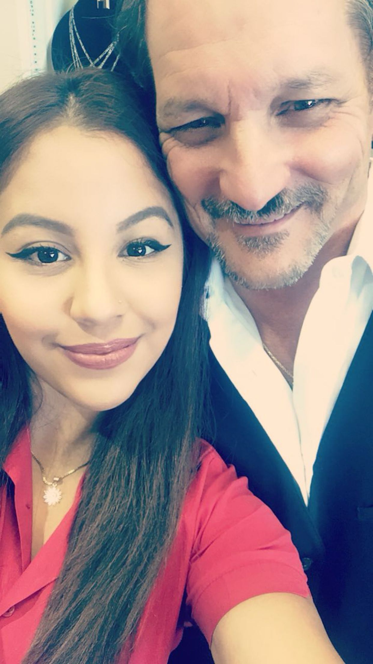 Two smiling people posing closely together indoors, one in red and one in a dark blazer.
