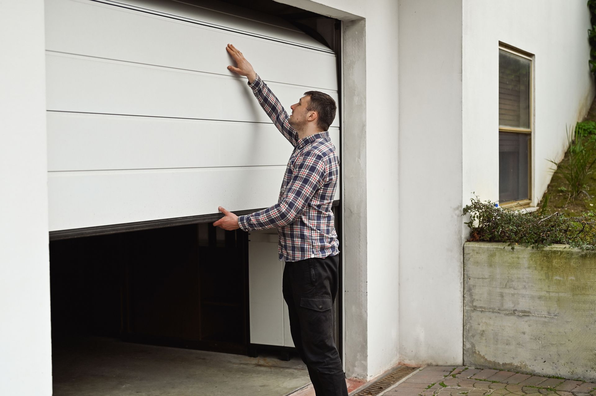 Man inspecting a white garage door. He wears a plaid shirt and black pants, and the setting is outside a house.