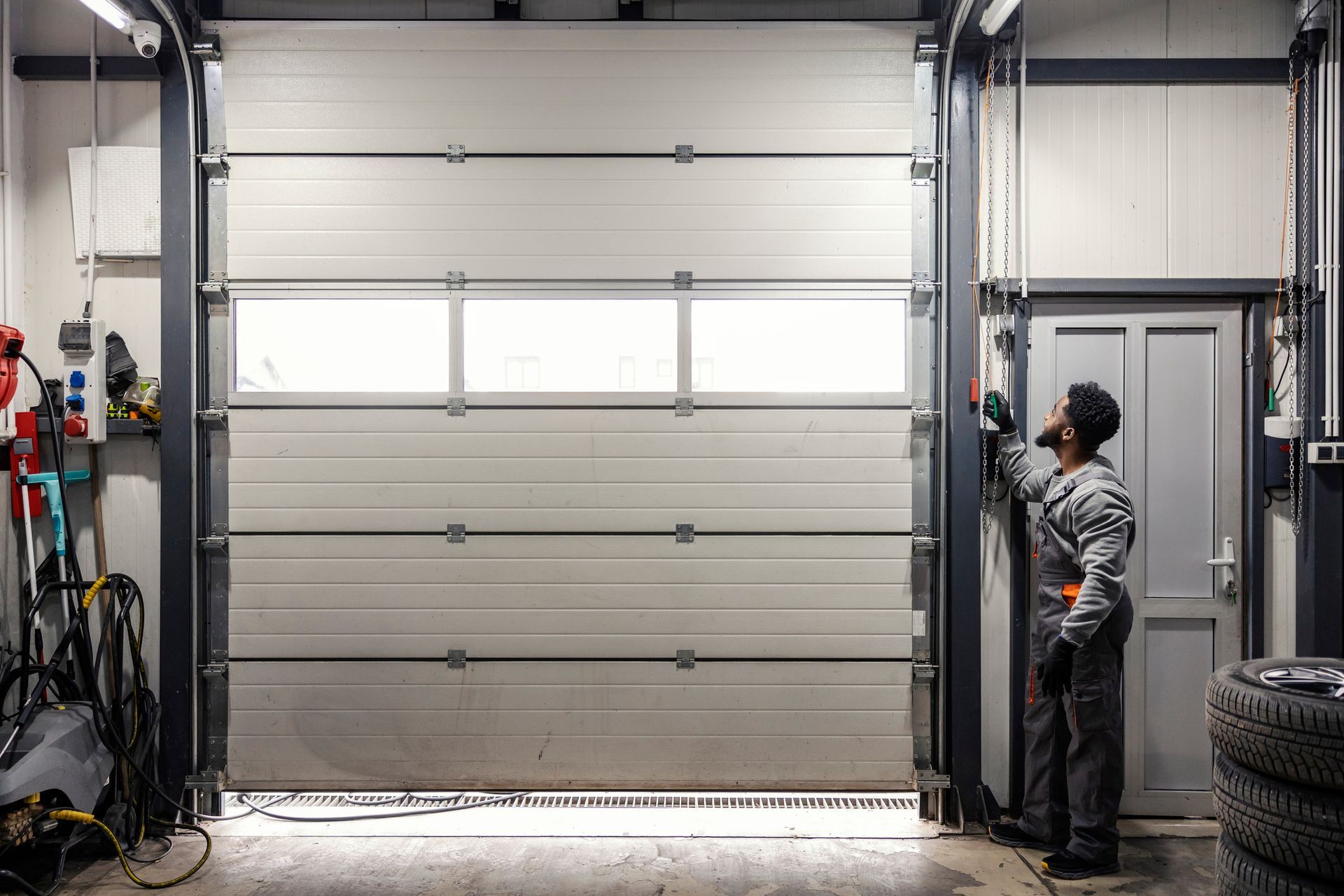 A mechanic in gray overalls inspects a closed white garage door. The setting is an industrial garage.