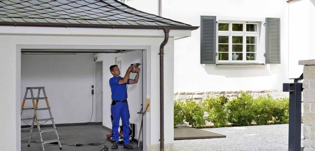 A man in blue overalls working on the inside wall of a white garage. There is a ladder in the garage.